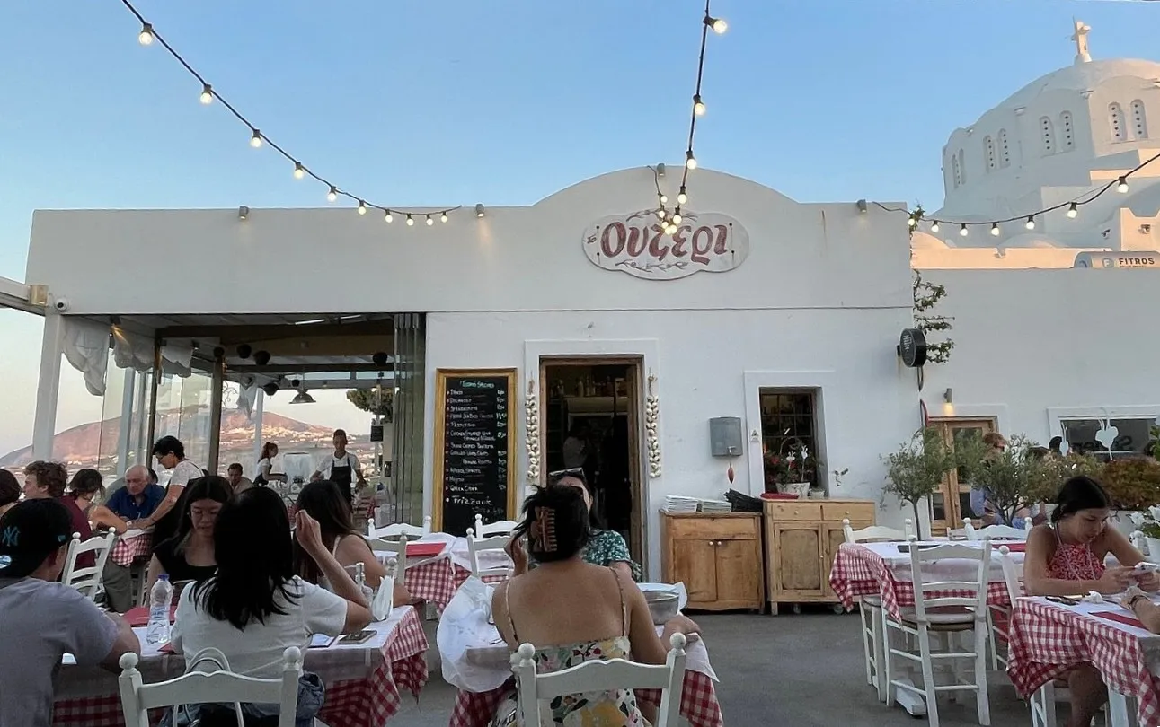 Guests dining at red-and-white checked tables on the Ouzeri terrace at dusk, under string lights, with the white dome of the Fira cathedral behind